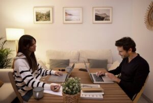 Two people working on laptops at a home table, with decorative pictures on the wall.