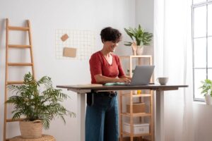 Woman smiling while using laptop at a standing desk in a bright home office with plants.