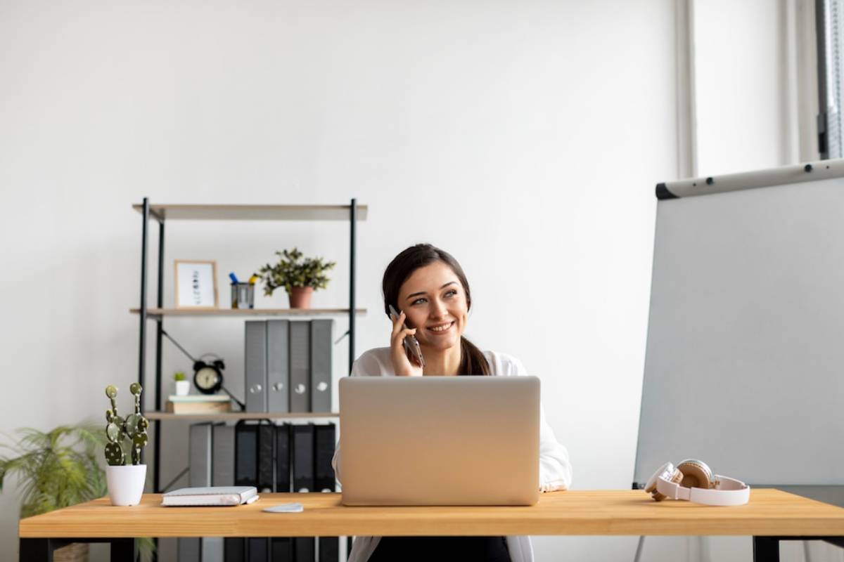 A smiling woman using a laptop and talking on a mobile phone in a modern office.