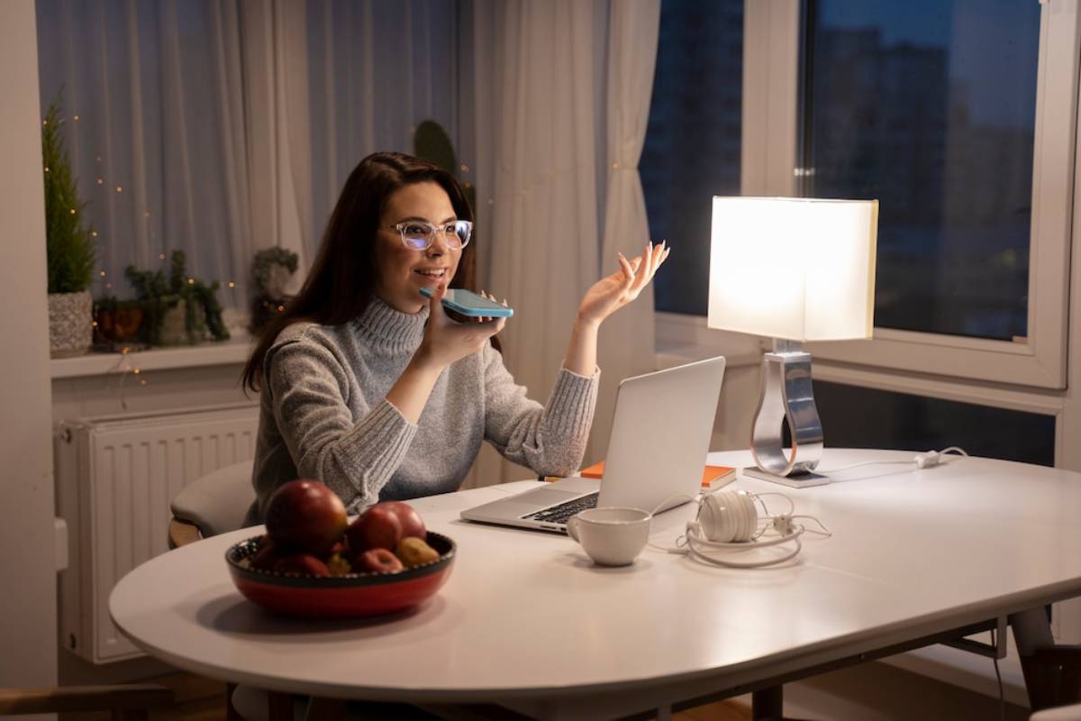Woman using a laptop and speaking into a smartphone in a cozy, well-lit room at night.
