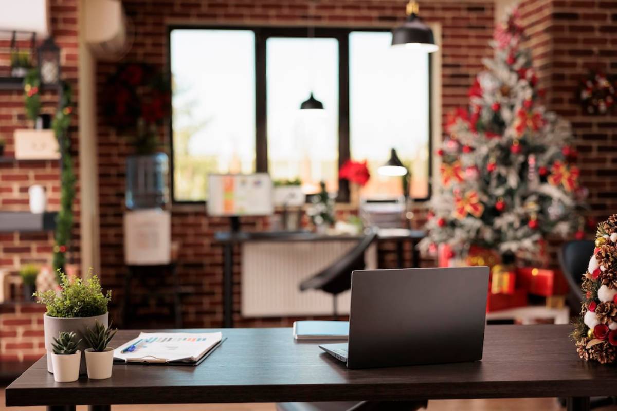A cosy office decorated for Christmas, featuring a laptop on a desk, with large windows and a festive tree in the background.