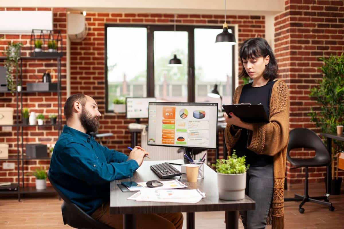 Two colleagues discussing work in a modern office with exposed brick walls and indoor plants.