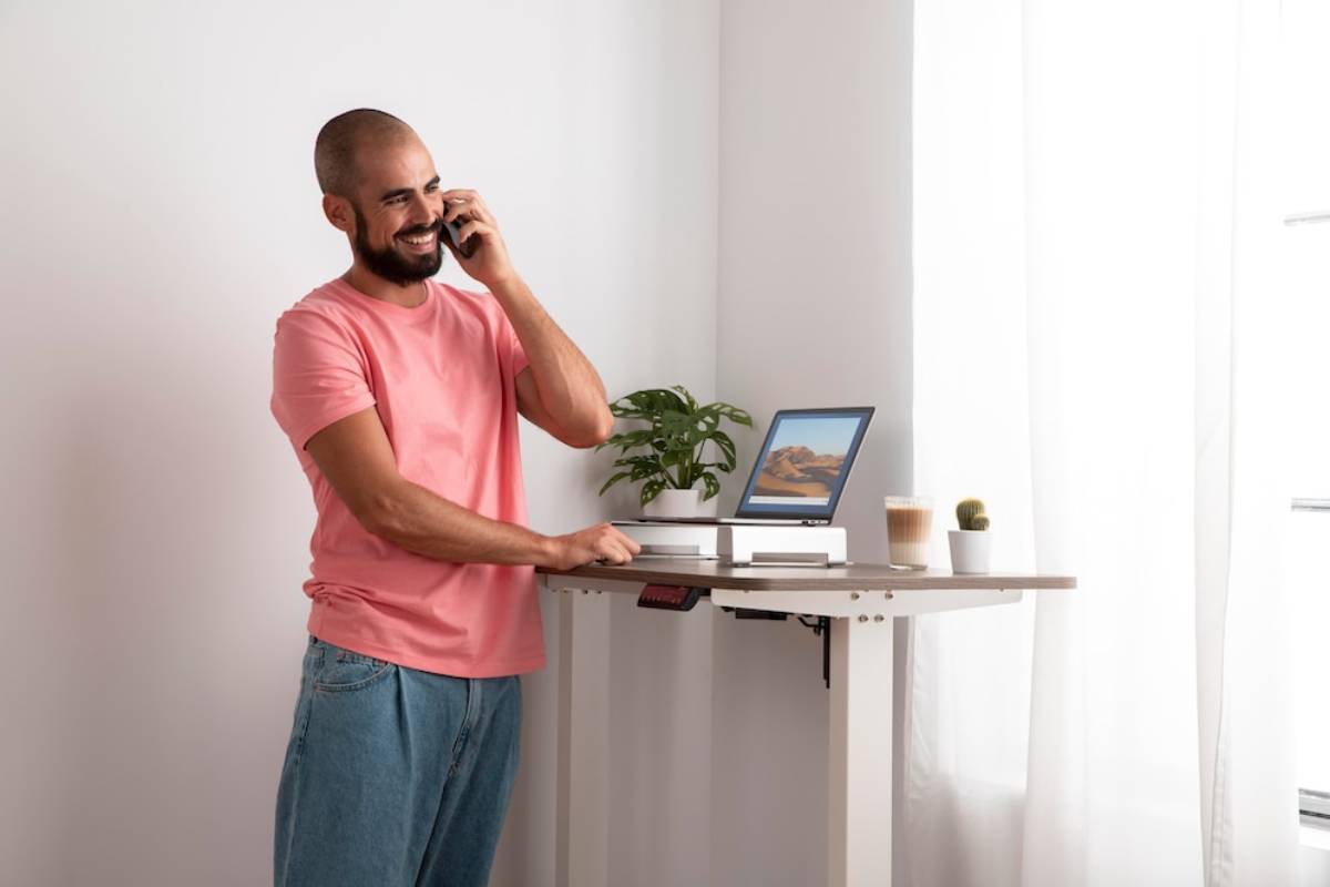 A man smiling while talking on the phone, standing at a standing desk with a laptop and a plant.