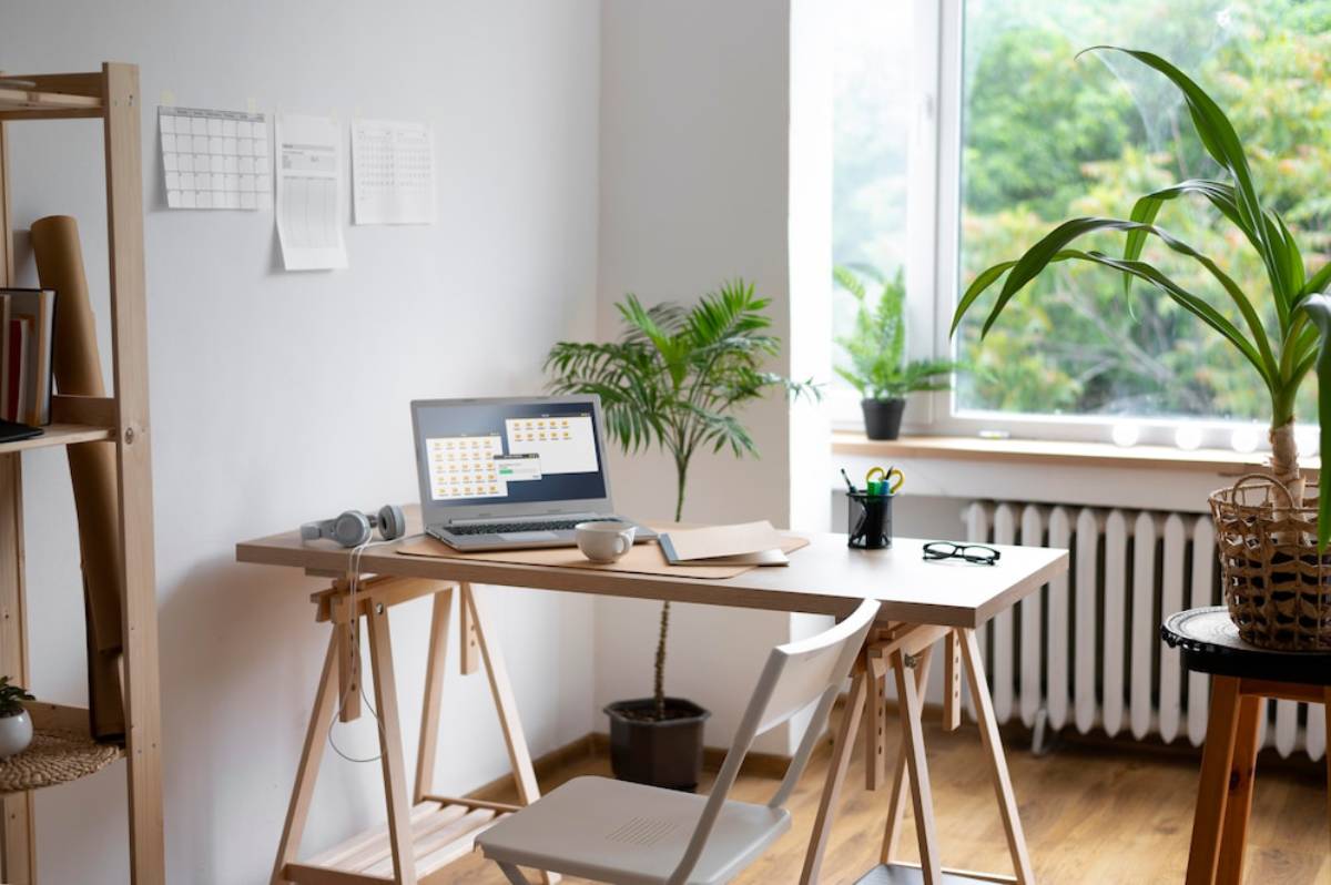 A bright home office with a laptop on a wooden desk, flanked by green plants and natural light from a window.