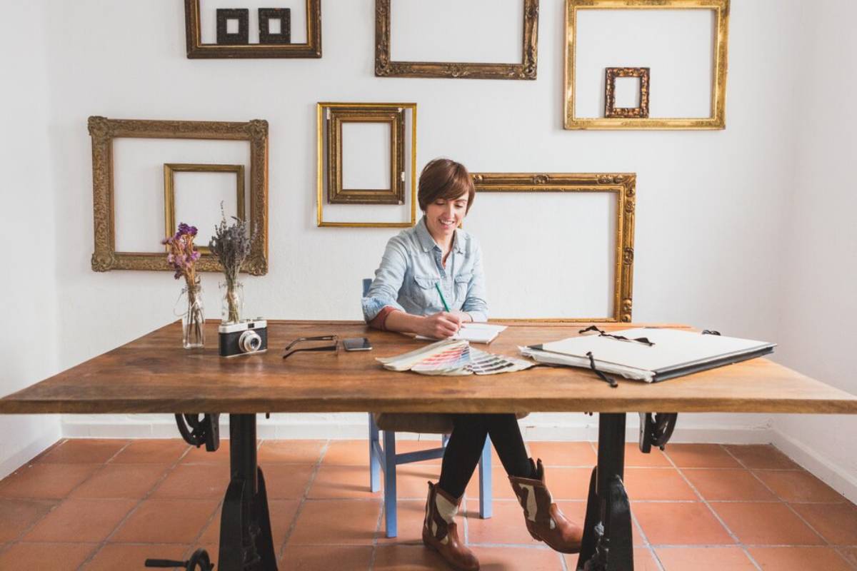 A woman sits at a large wooden table, writing in a notebook, surrounded by vintage picture frames on a white wall.