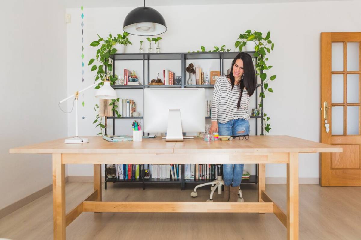 Woman standing in a well-lit home office with plants, shelving, and a wooden desk.