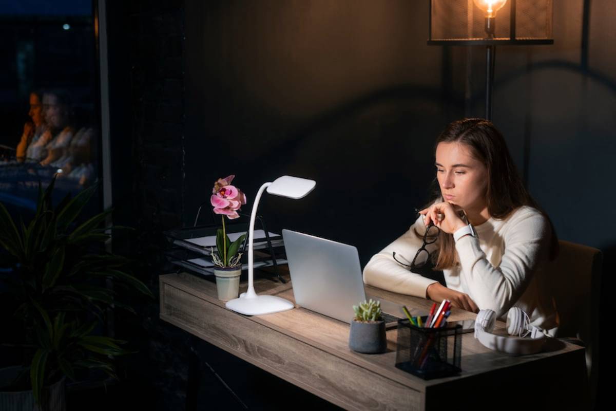 Woman working at night, focused on laptop in dimly lit room with desk lamp and plants.