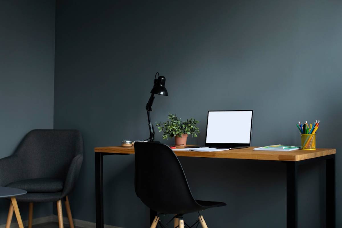A modern home office setup with a wooden desk, black chair, computer screen, desk lamp, and potted plant against a dark grey wall.