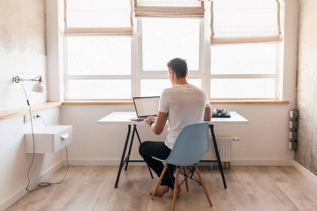 Man sitting at a simple desk with a laptop in a bright, sunlit room.