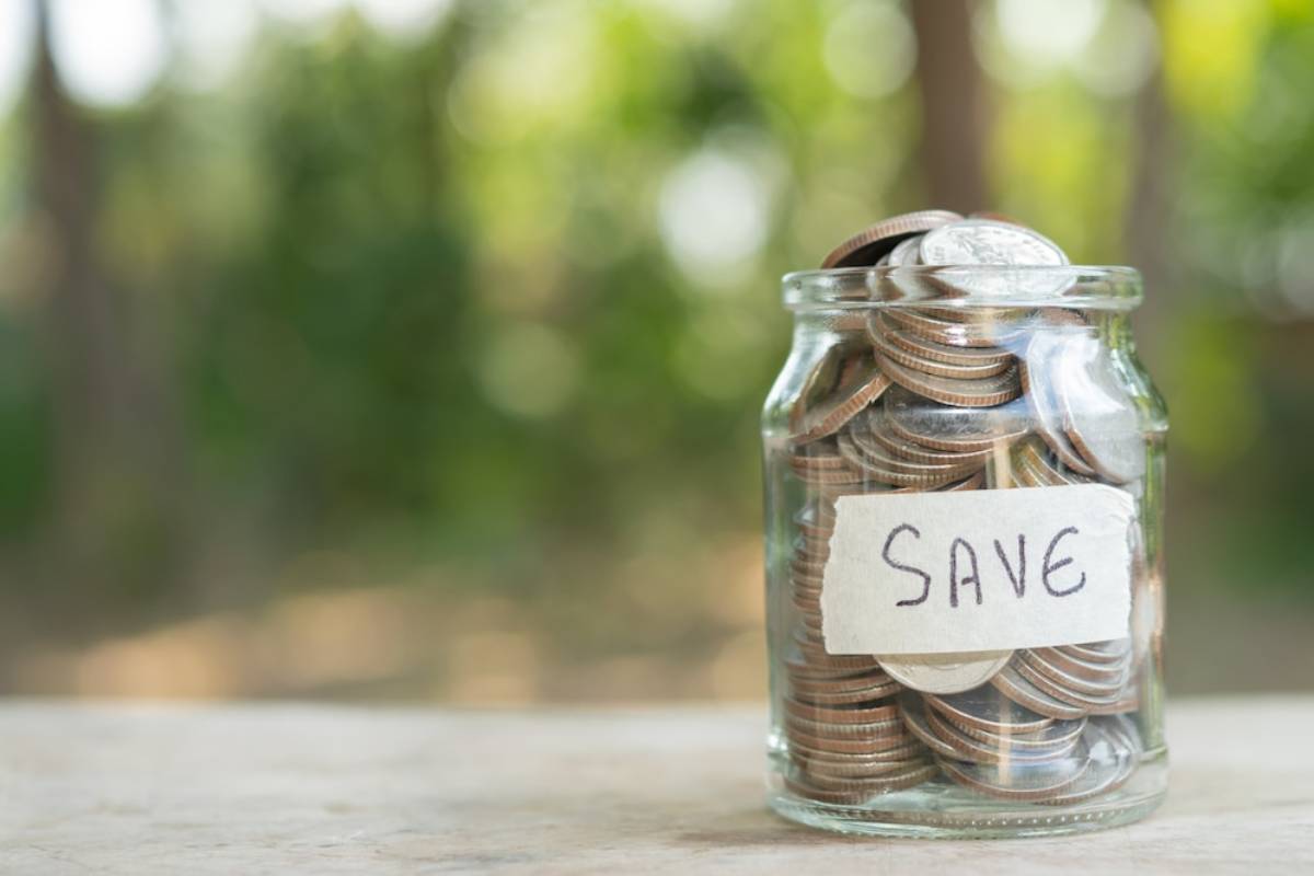 Glass jar filled with coins and labelled save on a wooden surface against a blurred green background.