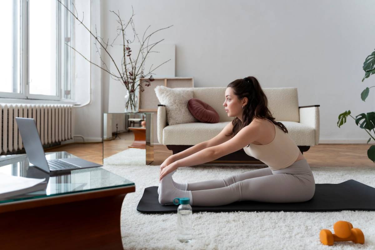 Woman stretching on a yoga mat at home, with a laptop and dumbbells nearby.