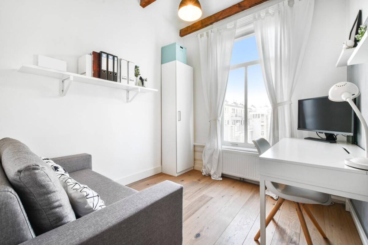 Bright, modern home office with grey sofa, white desk, chair, storage shelves, and large window with sheer curtains.