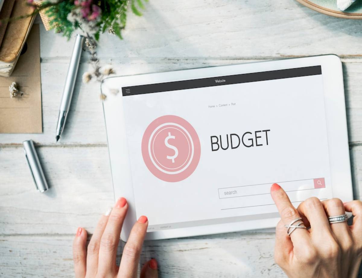 Hands pointing to a tablet displaying a budget planning screen on a wooden desk with stationery and flowers.