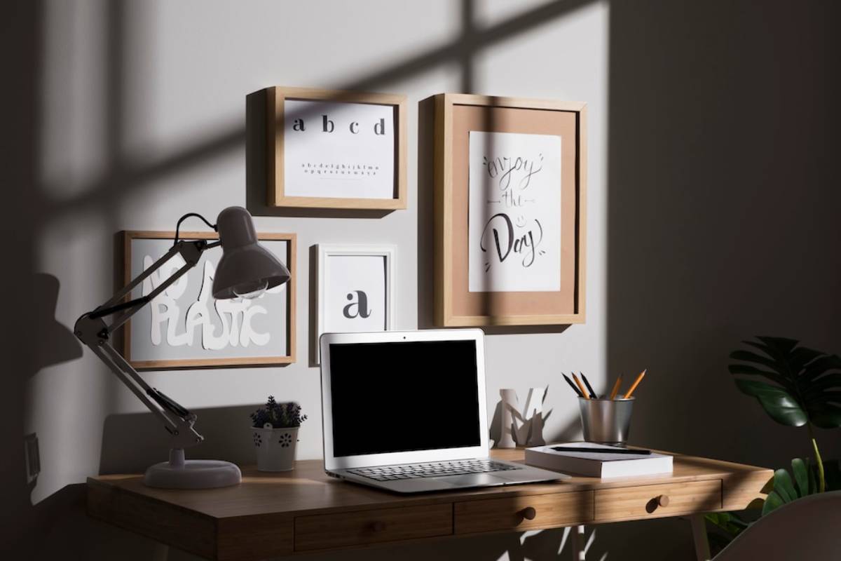 A modern home office with a laptop, desk lamp, and framed inspirational artworks under natural sunlight.