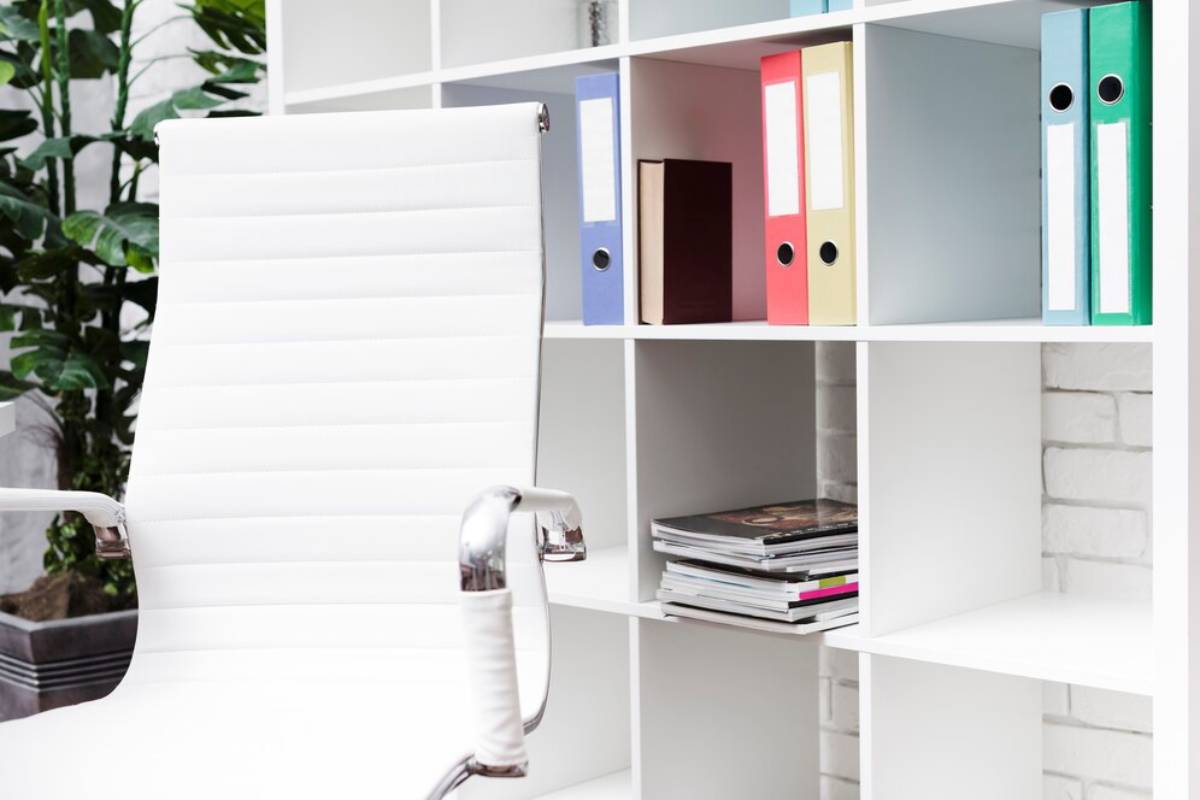 A modern office with a white chair, colorful binders on shelves, and indoor plants.