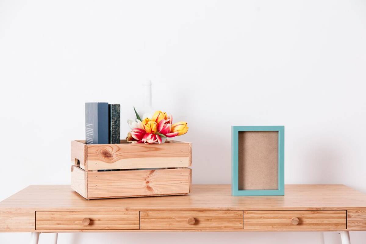 Wooden table with books, flowers in vase, and empty turquoise picture frame against a white background.
