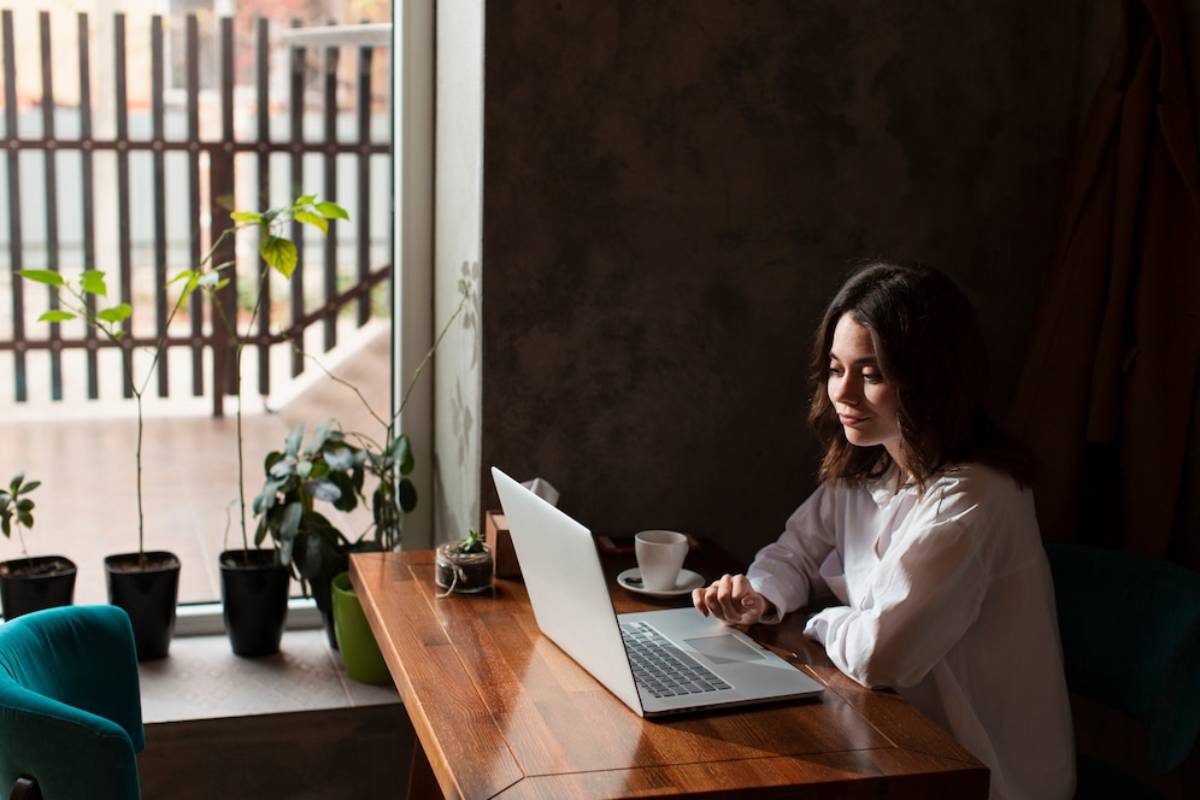Woman working on laptop at a wooden table by a window, surrounded by potted plants, in a warmly lit room.