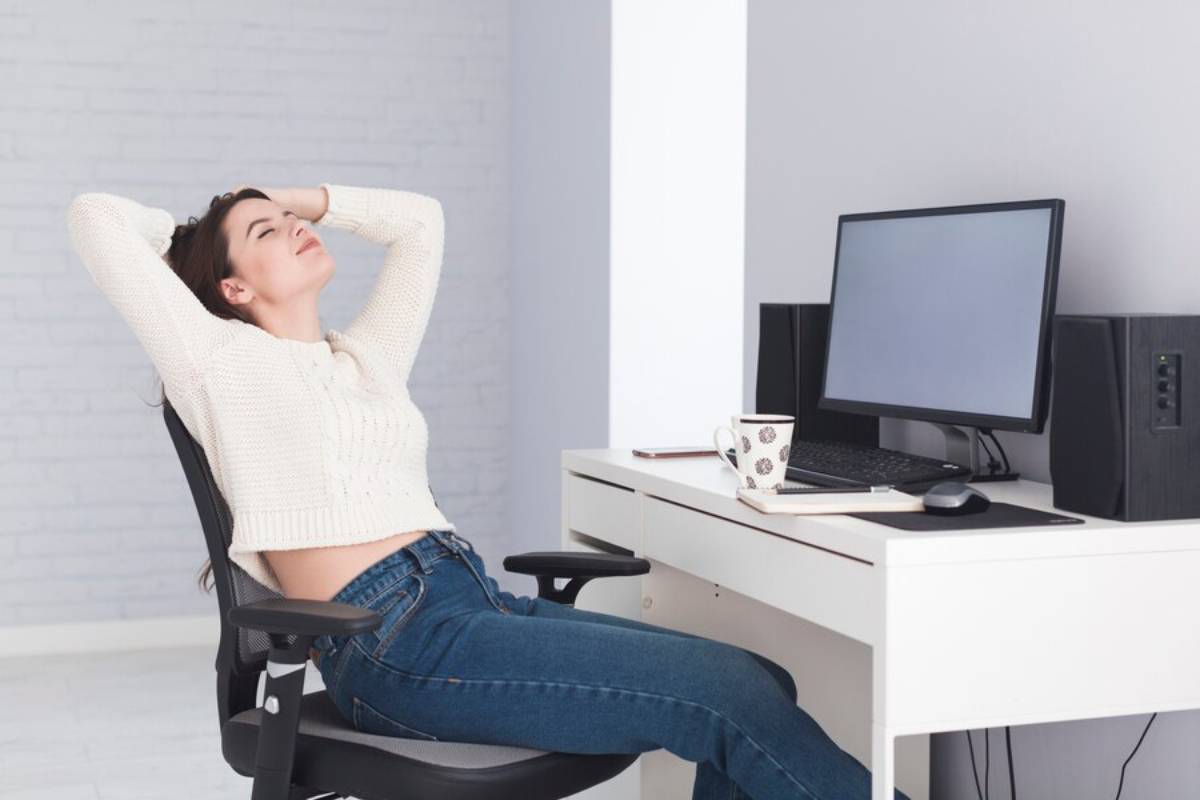 A woman in a white sweater and jeans leans back in an office chair, hands behind her head, in front of a desk with a computer, mug, phone, and speakers.