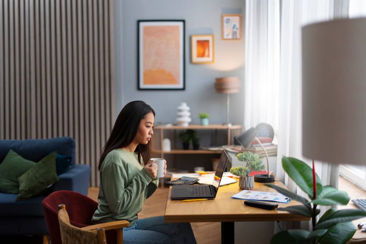 A woman working on a laptop at a home office desk, holding a mug.