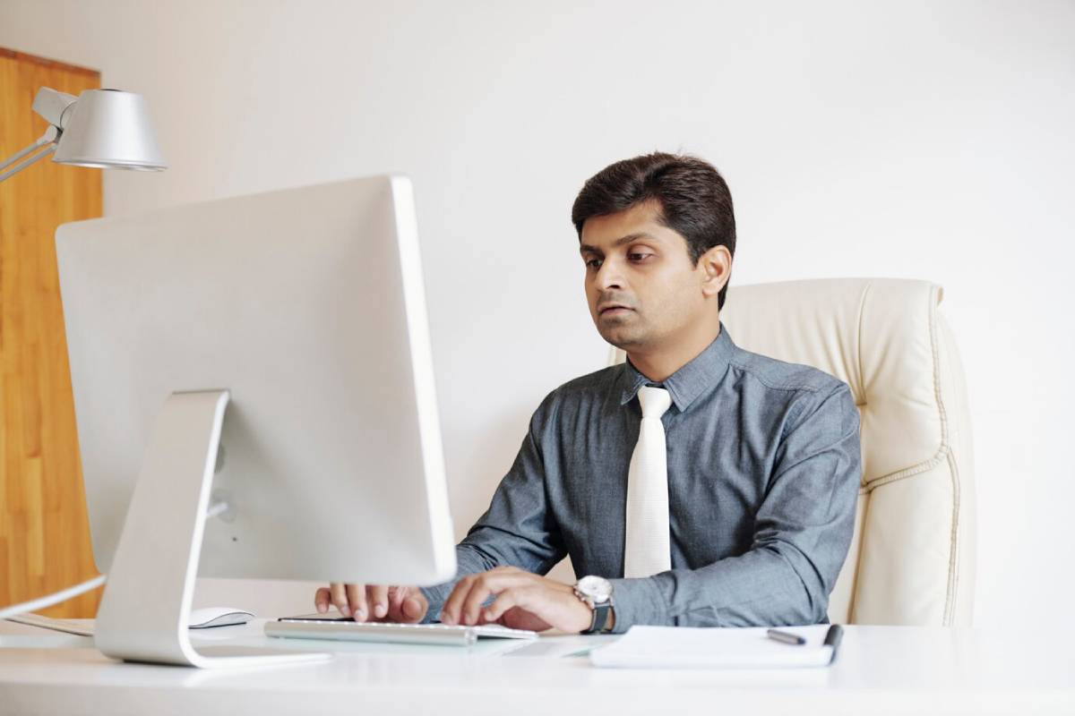 A man in a shirt and tie sits at a desk, working on a computer in a professional office setting.