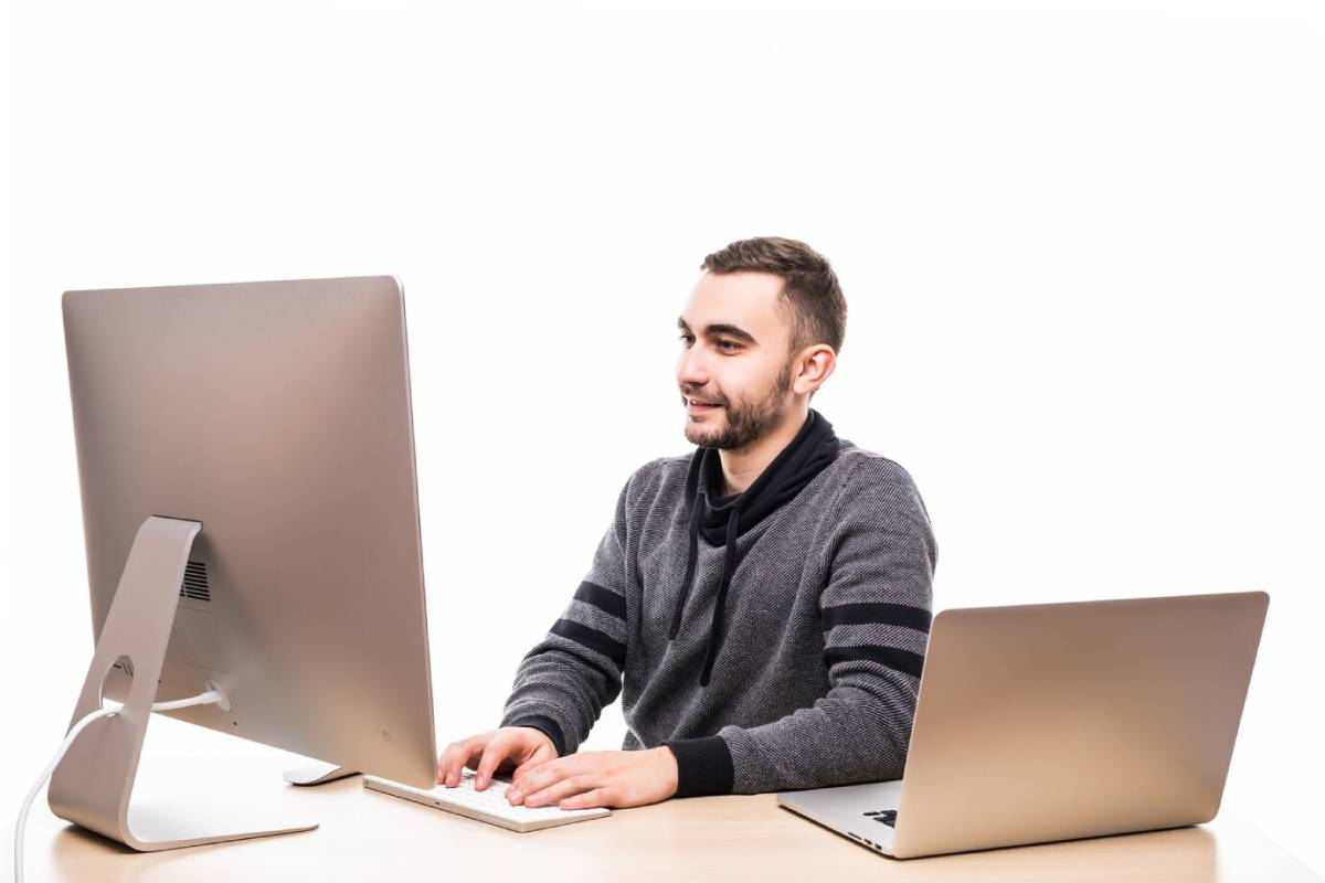  A man sits at a desk, working on a computer and a laptop, focused on his tasks.