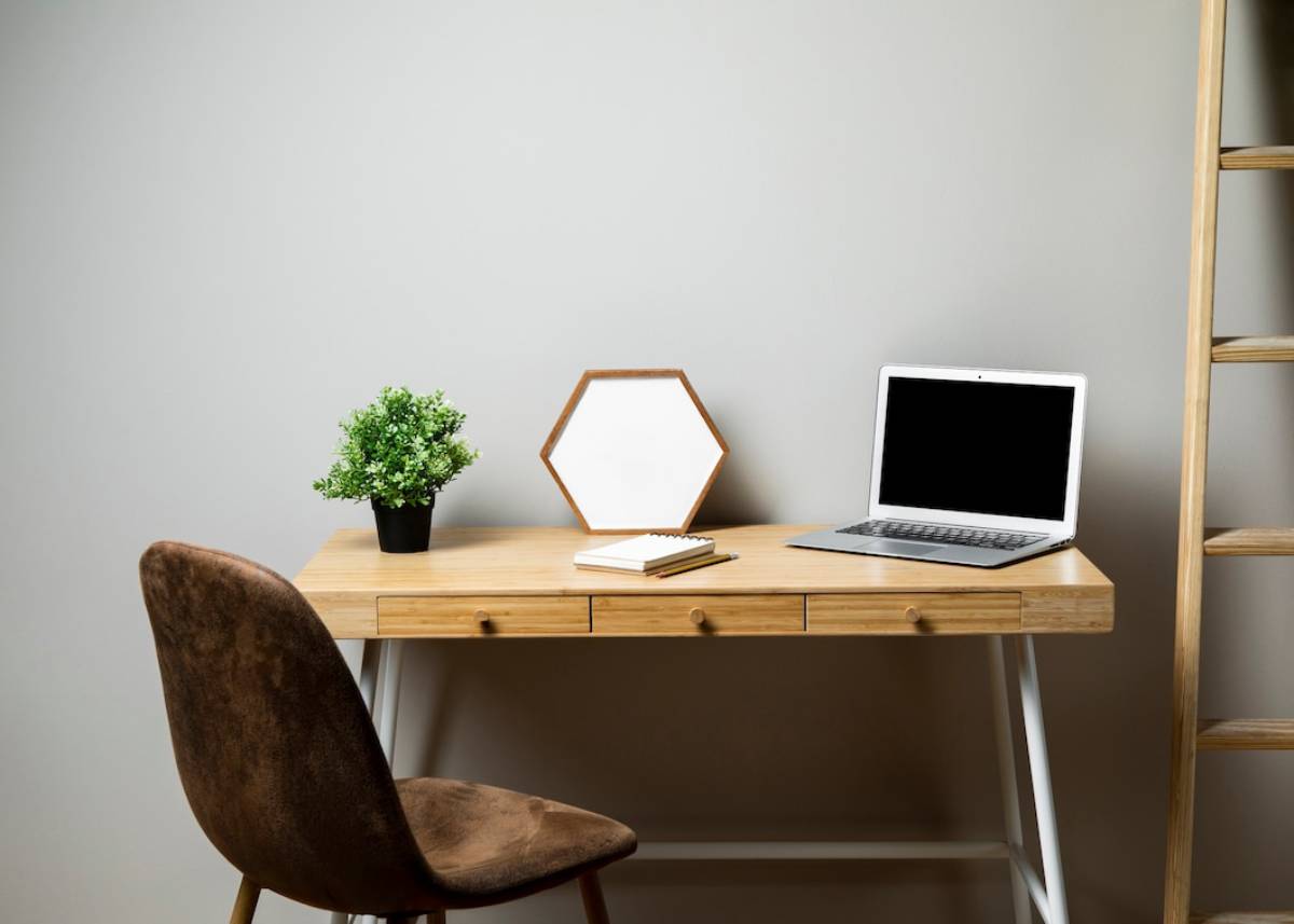 A laptop sits on a wooden desk beside a chair, creating a simple workspace setup.
 