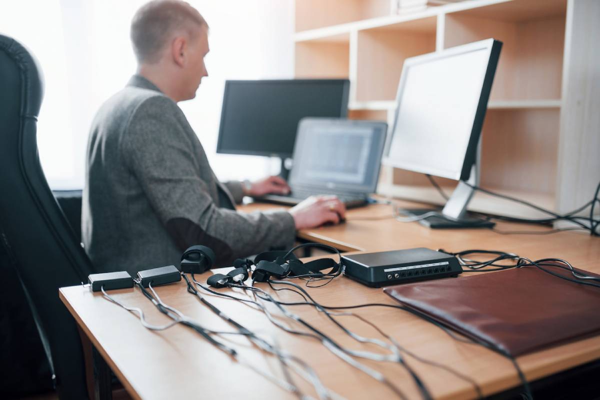A man seated at a desk with a computer, surrounded by numerous tangled wires.