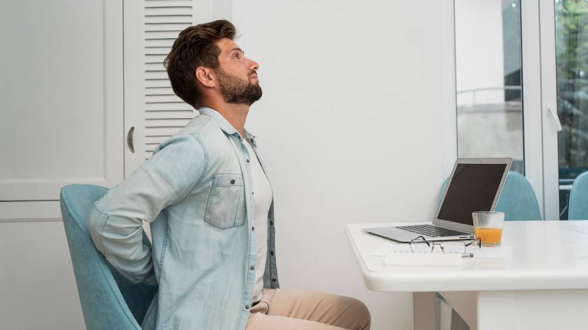 Man sitting at a desk, visibly uncomfortable, holding his lower back due to pain. 