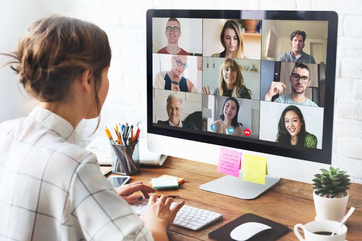 A woman sits at a desk, viewing a video call with a group of people on her computer screen.