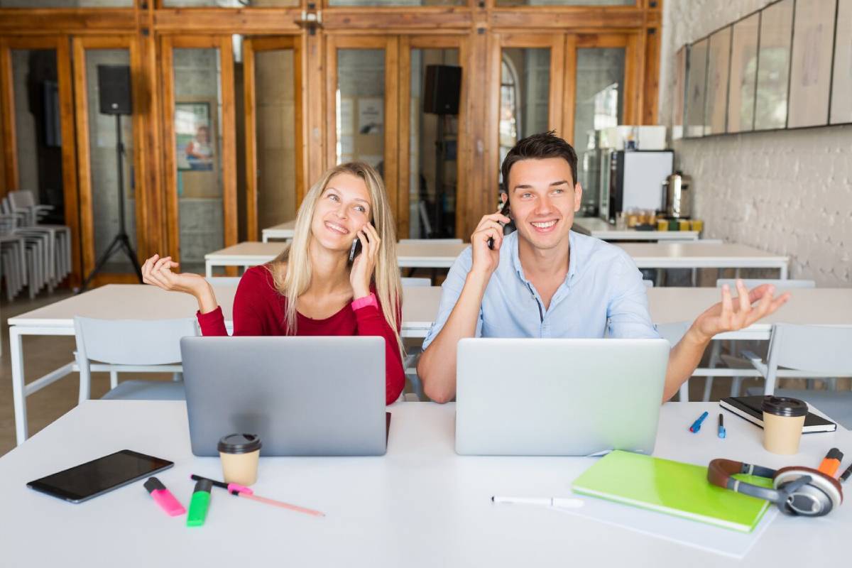 Two people at a table with laptops, engaged in conversation while talking on their phones. 