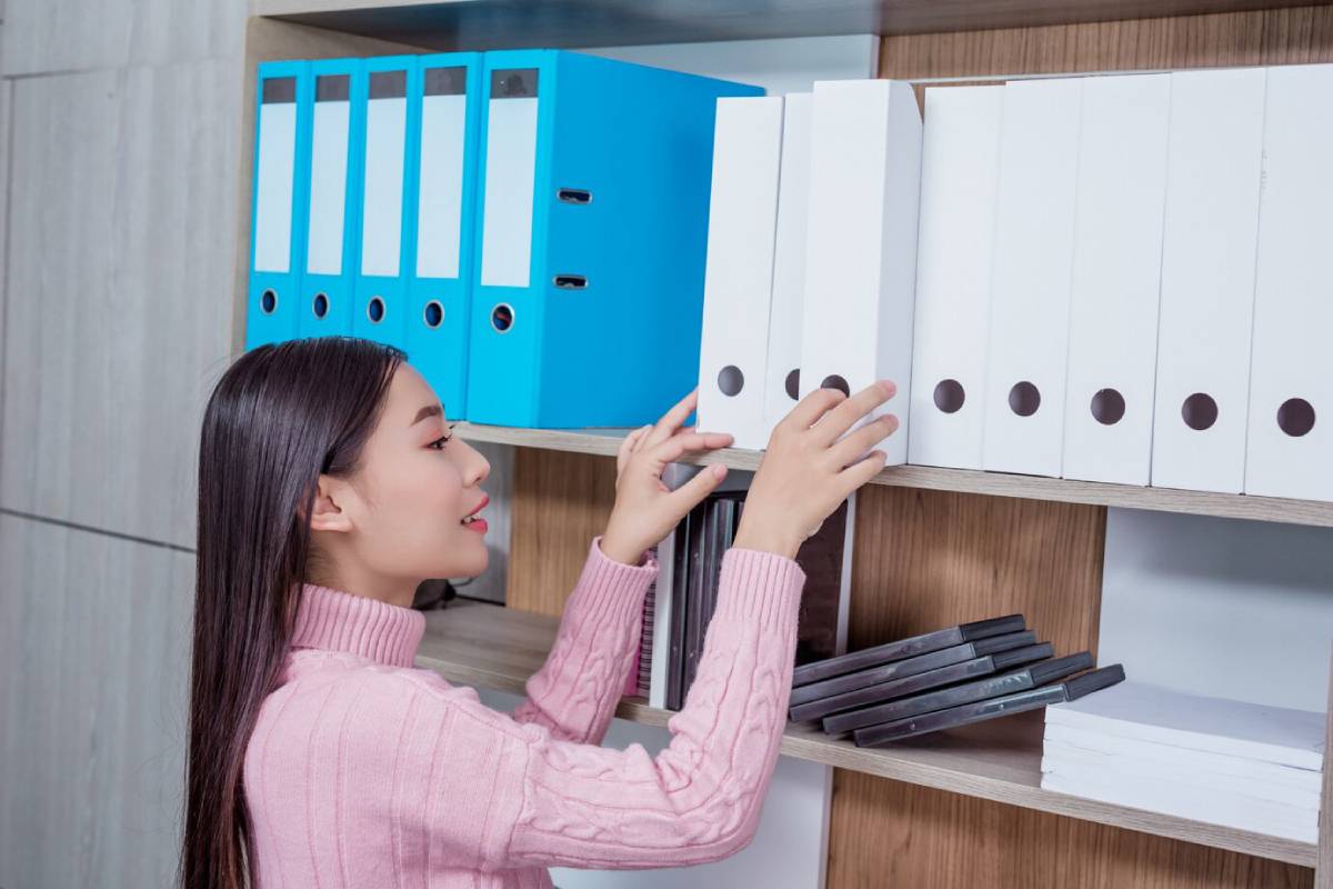 A woman holds a file folder while standing in front of a shelf filled with various documents and folders. 